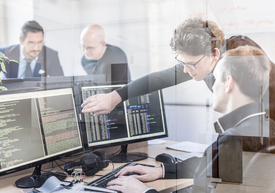 Офис обстановка. Колега показва на друг, седящ на бюро, нещо на два големи монитора.