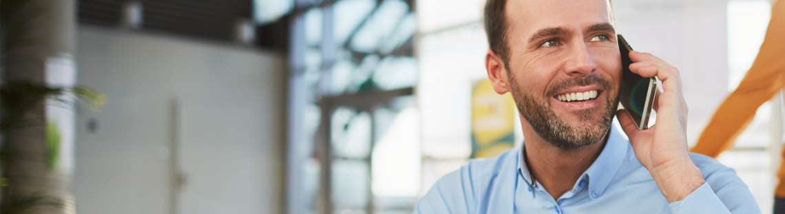 Млад човек, седнал във фотьойл в офисна среда, говори по телефон с усмивка.