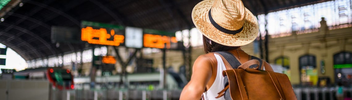 Жена, носеща шапка и раница, стои пред информационно табло на летище.