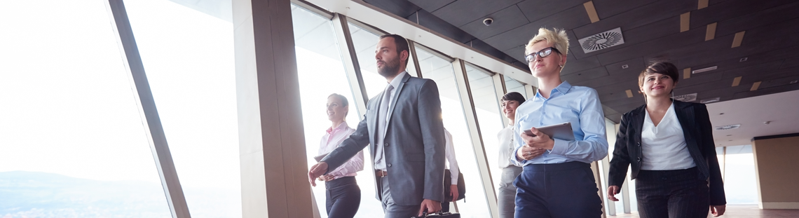 Бизнес екип от шест души върви през коридор на бизнес сграда с остъклена стена с гледка към града.