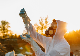 Egy maszkot és kesztyűt viselő személy, aki egy főzőpoharat tart a kezében.
