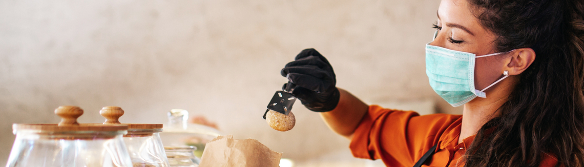 Egy maszkot és kesztyűt viselő nő egy zacskó ételt tart a kezében a pékségben