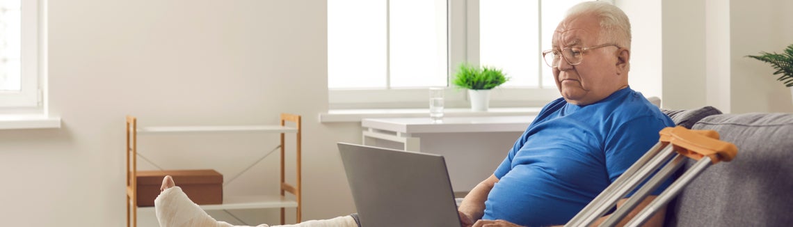 Starší muž se zlomenou nohou sedící na pohovce a používající notebook