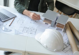 На работен плот има чертеж, компютър и предпазна каска, а ръка показва макет на къща.