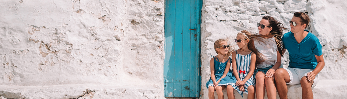 Младо семейство с две дъщери седят пред бяла стена със синя врата.
