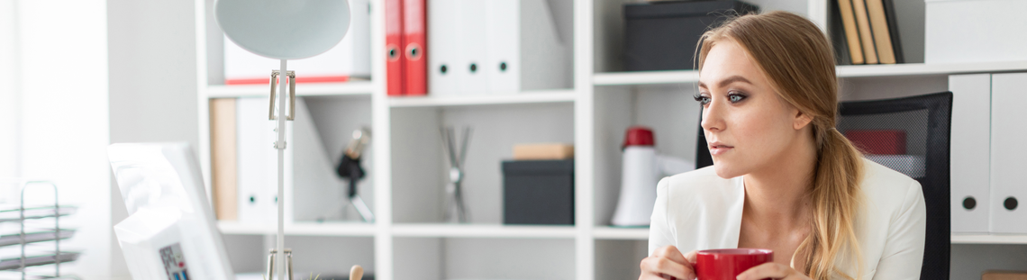 Жена зад бяло офис бюро пие кафе в червена чаша и гледа замислено в монитора.