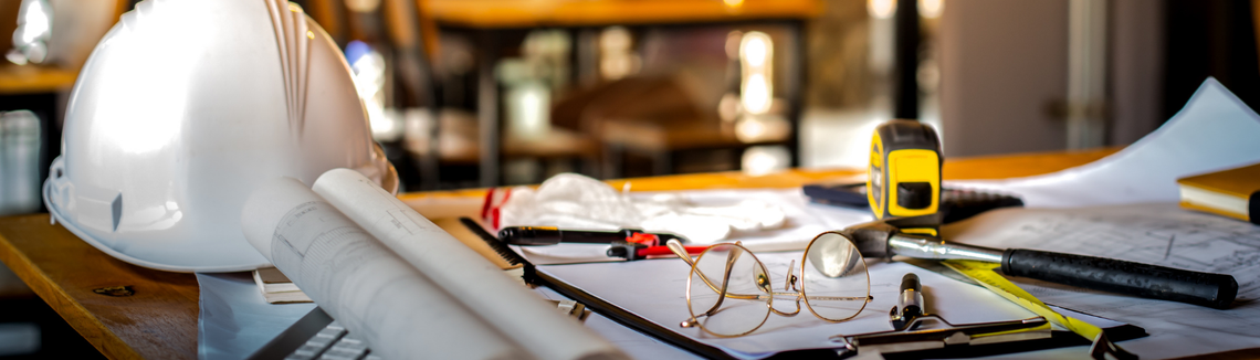 Проектантска маса със свит на руло чертеж, предпазна каска, ролетка, очила и документи.