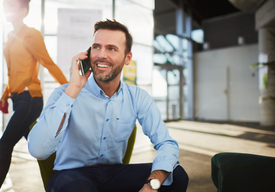 Млад човек, седнал във фотьойл в офисна среда, говори по телефон с усмивка.
