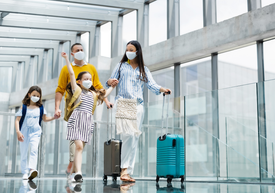 Младо семейство с две момиченца с маски на лицата, които бутат куфарите си из летище.