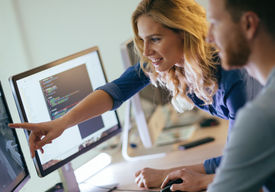 Жена показва на свой колега, който стои на бюро с два големи монитора, нещо на единия екран.