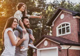 Egy fiatal pár, aki egy gyermeket visz a vállán egy ház előtt.