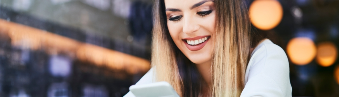 Млада жена усмихнато гледа екрана на мобилния си телефон.