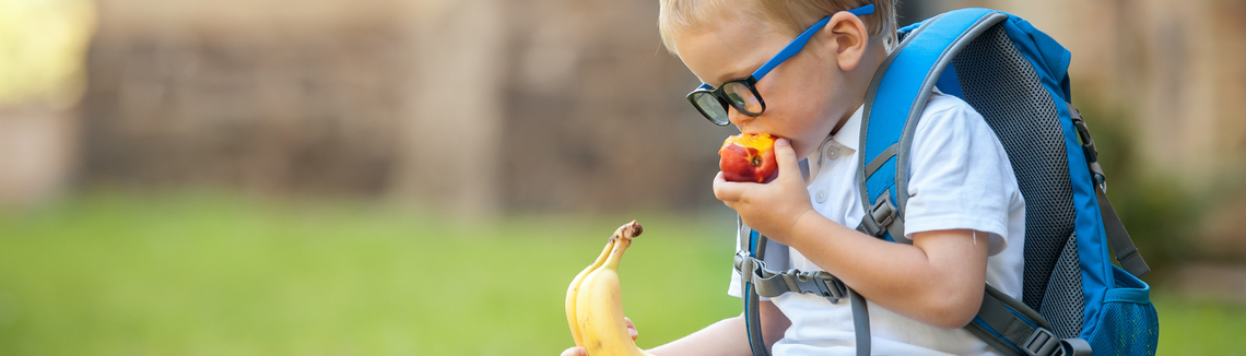 chłopiec z niebieskim plecakiem je jabłko i banana na przerwie w szkole
