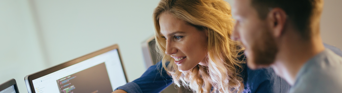 Жена показва на свой колега, който стои на бюро с два големи монитора, нещо на единия екран.