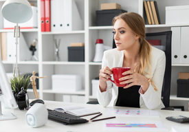Жена зад бяло офис бюро пие кафе в червена чаша и гледа замислено в монитора.