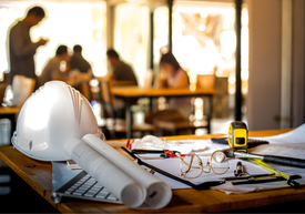 Проектантска маса със свит на руло чертеж, предпазна каска, ролетка, очила и документи.