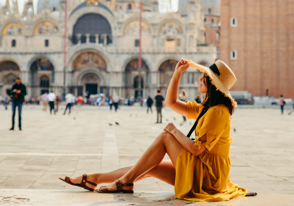 Egy nő ül a földön egy épület előtt sárga ruhában és fogja a kalapját.