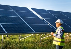 Мъж в светлотразителна жилетка и каска стои пред соларни панели