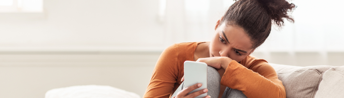 Момиче, седнало на дивани със свити пред себе си крака, гледа умишлено в телефона си.