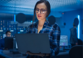 Млада жена в синя карирана риза държи лаптоп, а на заден синкав фон се вижда офис обстановка.