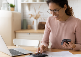 Uśmiechnięta kobieta w okularach ssedzi przed komputerem i liczy na kalkulatorze.