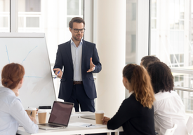 Un bărbat în costum stă în fața unei table albe cu oameni în jurul său