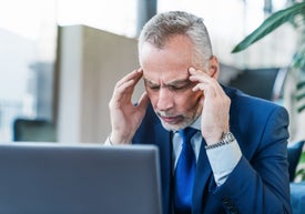 Мъж в костюм докосва главата си видимо разтревожен над лаптопа си.