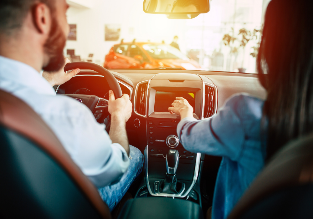 Кадър, сниман от задната седалка на кола, отпред седят мъж и жена, която показва нещо на таблото.