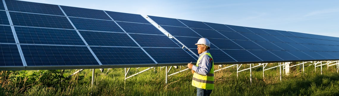 Мъж в светлотразителна жилетка и каска стои пред соларни панели.