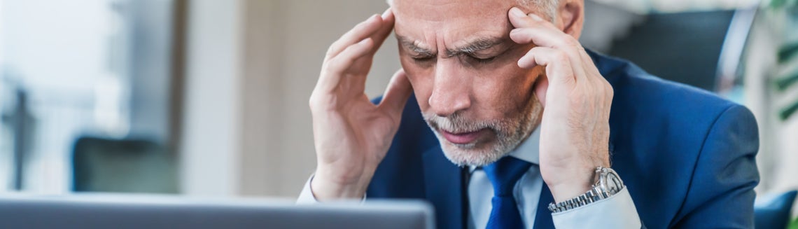 Мъж със затворени очи, който държи главата си, разтревожен над лаптопа си.