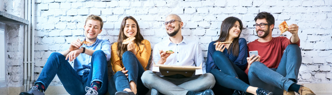 Skupina ľudí sedí na zemi a jedia pizzu