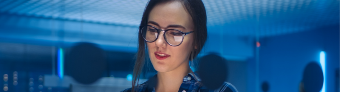 Млада жена в синя карирана риза държи лаптоп, а на заден синкав фон се вижда офис обстановка.