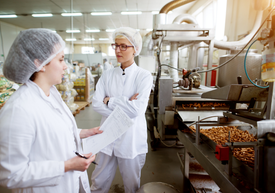 Žena v bielom plášti stojaca vedľa ženy v továrni