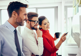 Трима офис служители усмихнати разглеждат листове, закачени на табло.