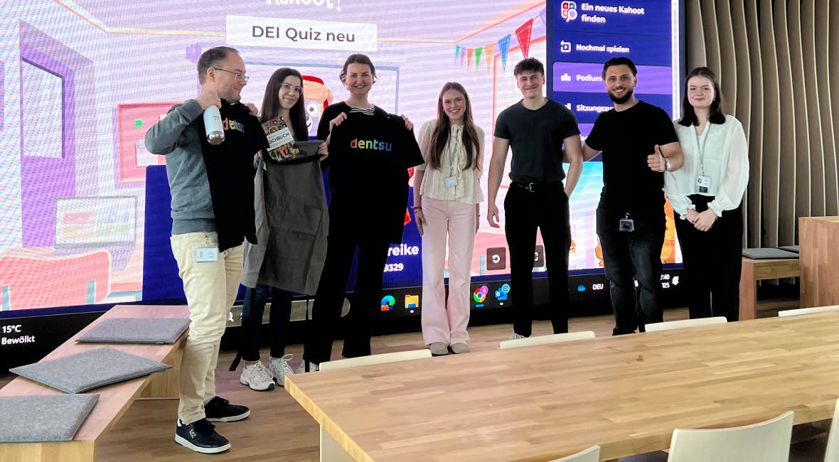 Gruppe von Mitarbeitenden steht vor großem Bildschirm mit Kahoot‑Quiz im Büro, einige halten Preise in den Händen.