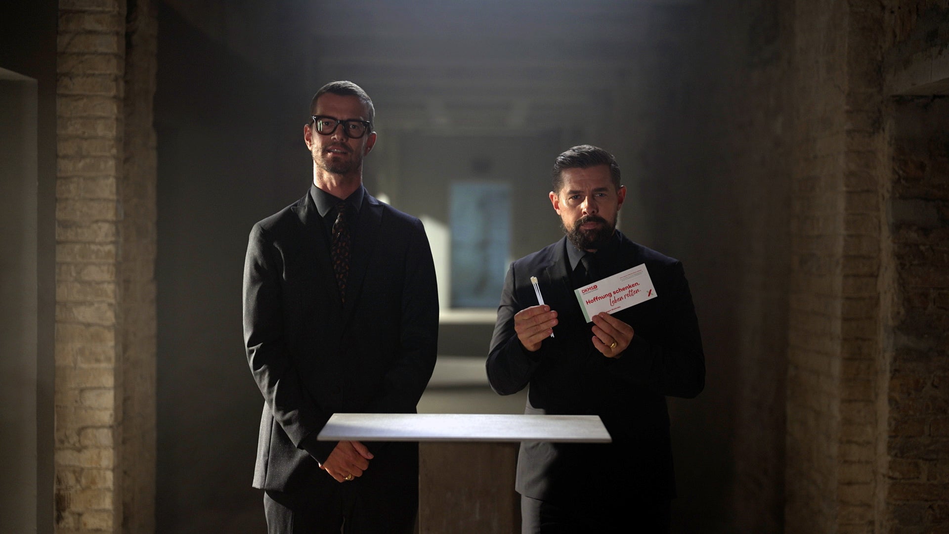 Zwei Männer in dunklen Anzügen stehen nebeneinander in einem schmalen, gemauerten Gang mit Backsteinwänden. Beide haben die Hände vor dem Körper gefaltet. Vor ihnen schwebt ein rechteckiges, weiß leuchtendes Objekt. Der Gang ist nach hinten perspektivisch verlängert und durch Lichtschächte beleuchtet.