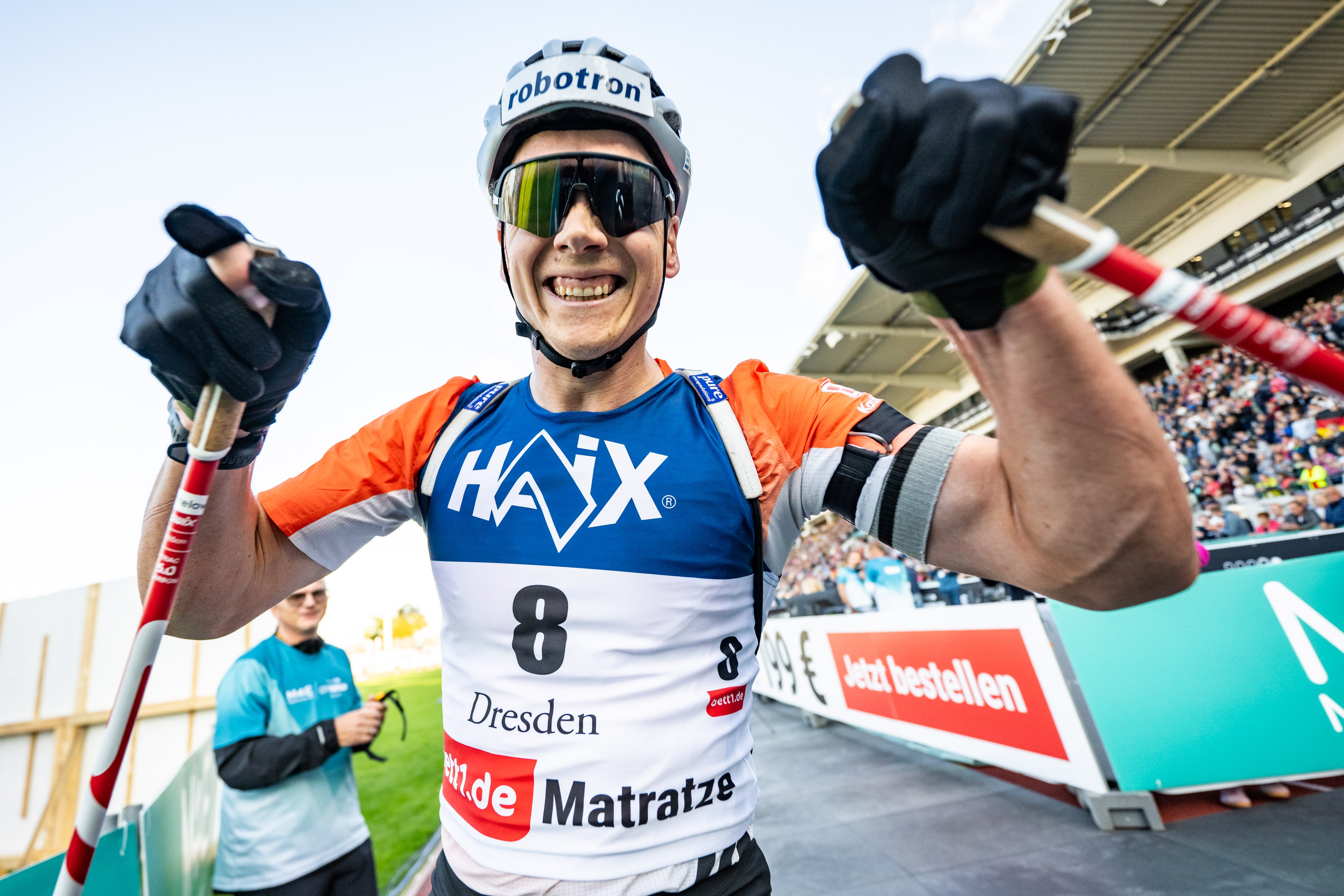 Ein Biathlon-Athlet lächelt in die Kamera, hinter ihm ist ein Publikum im Stadion zu sehen. 