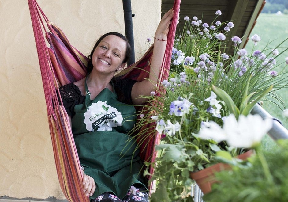 Eine Frau in einer grünen Schürze sitzt in einer Hängematte umgeben von Blumen