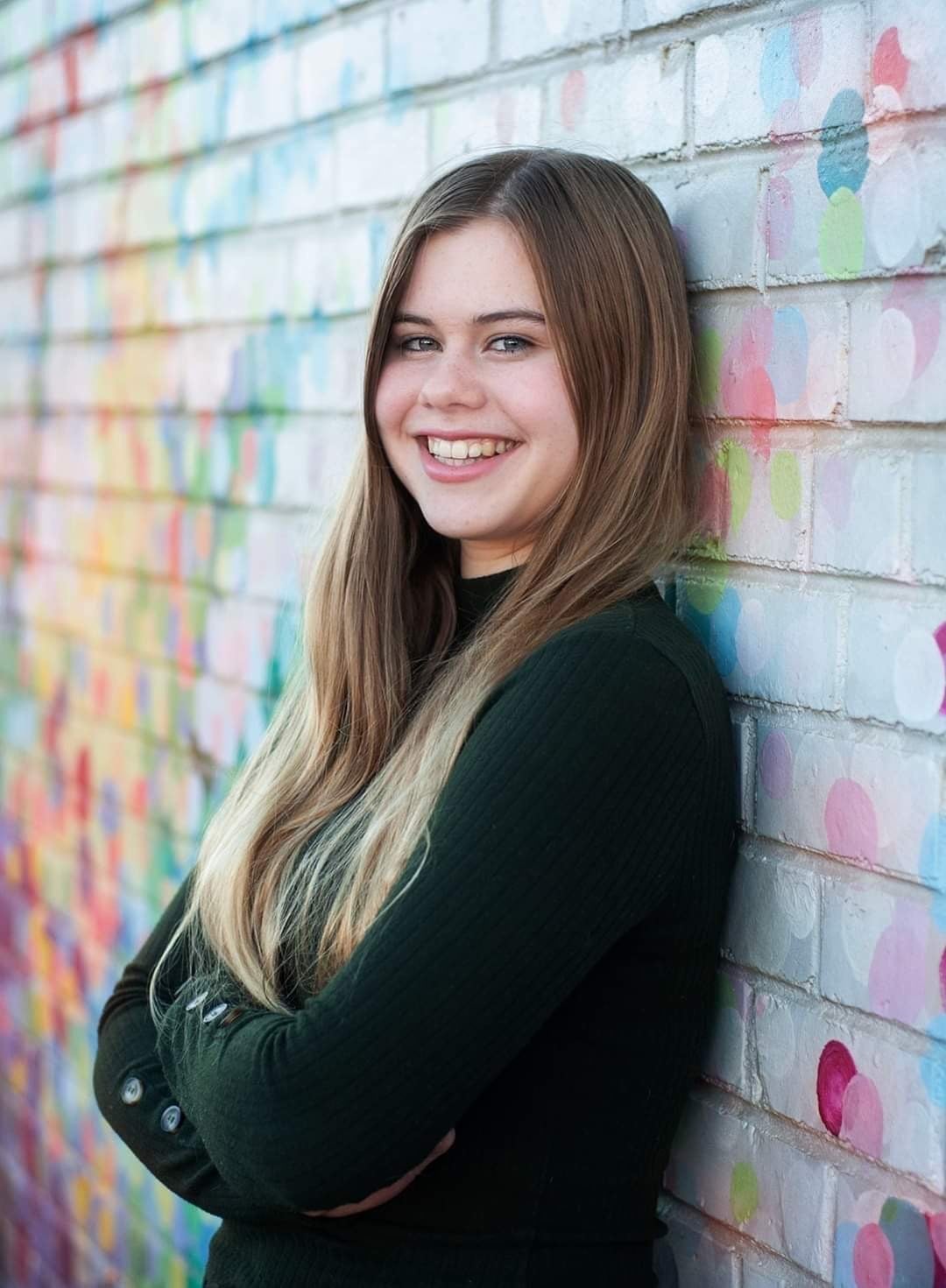 Eine Frau lehnt mit verschränkten Armen an einer bunten Mauer und lächelt.