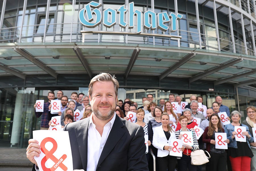 Eine Gruppe von Menschen steht vor einem Gebäude der Gothaer. Alle halten weiße Zettel mit dem Logo der DKMS hoch. 
