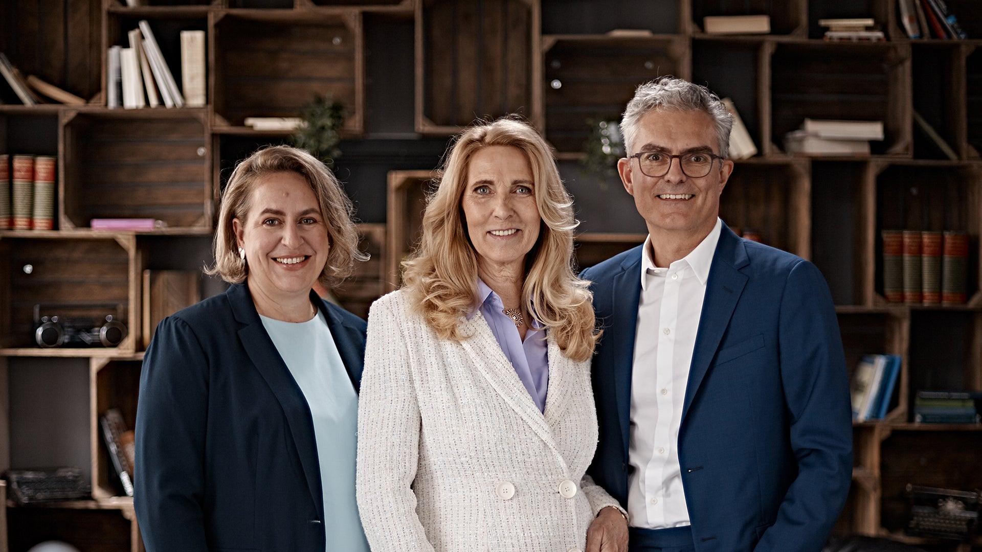 Porträt von Dr. Elke Neujahr, Dr. Deborah Buk und Stephan Schumacher im Business-Outfit vor einem Regal mit Büchern und Dekoration.