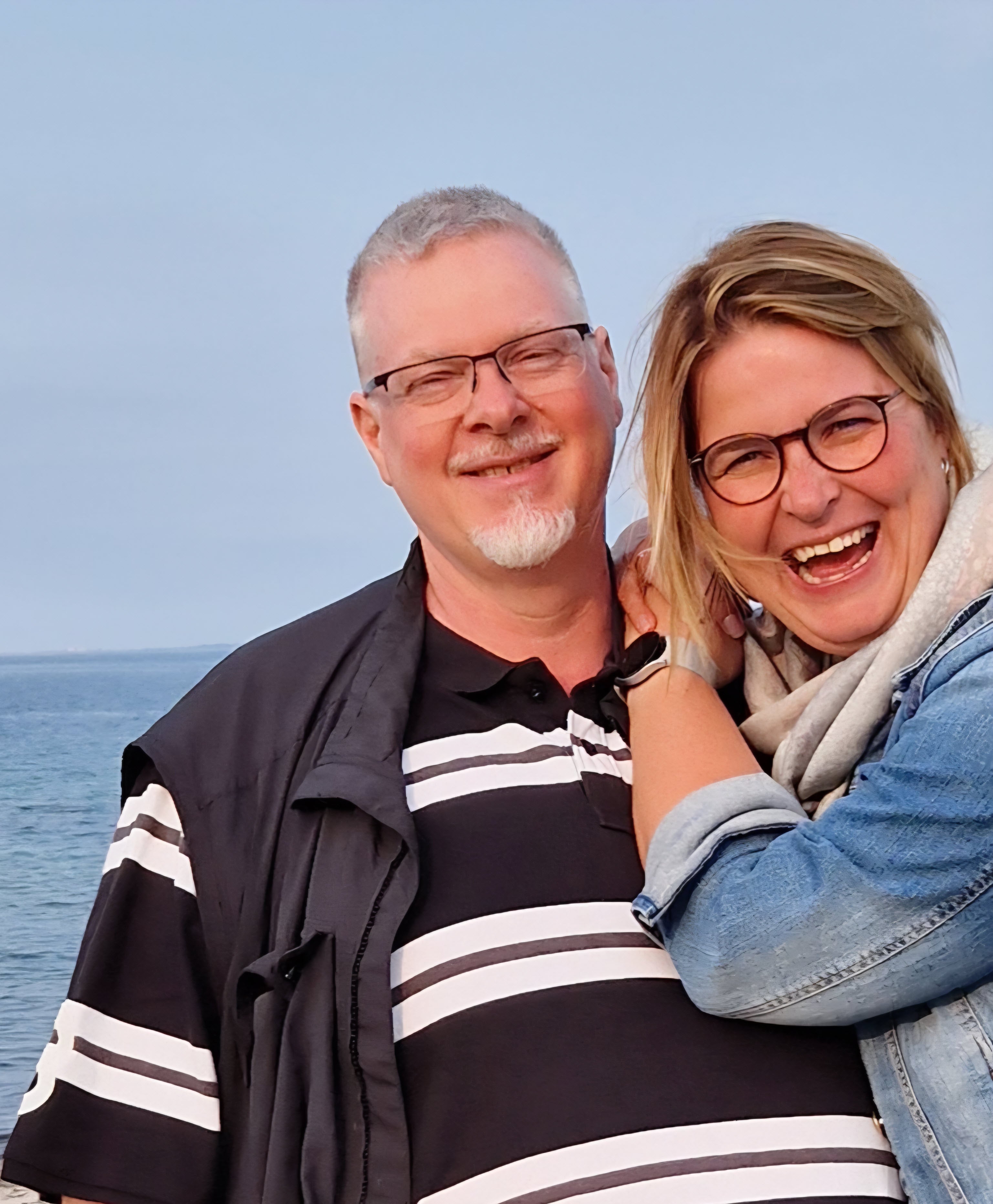 Patient Jürgen und seine Frau lachen in die Kamera. Hinter ihnen ist Wasser zu sehen