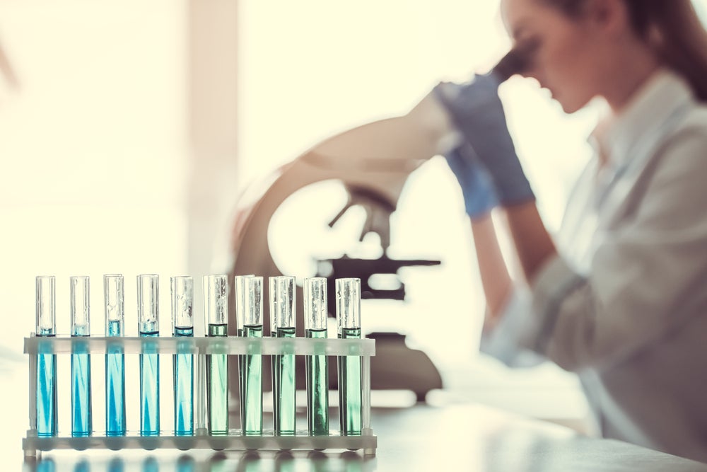 Eine Gruppe von Reagenzgläsern mit blauer Flüssigkeit, im Hintergrund eine Frau die in ein Mikroskop guckt