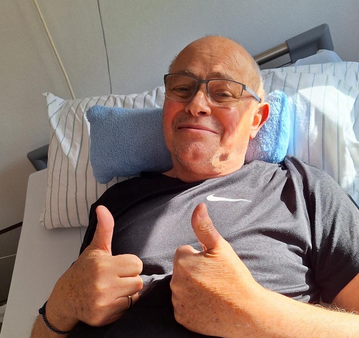 Ein Mann liegt in einem Krankenhausbett und zeigt mit beiden Daumen nach oben