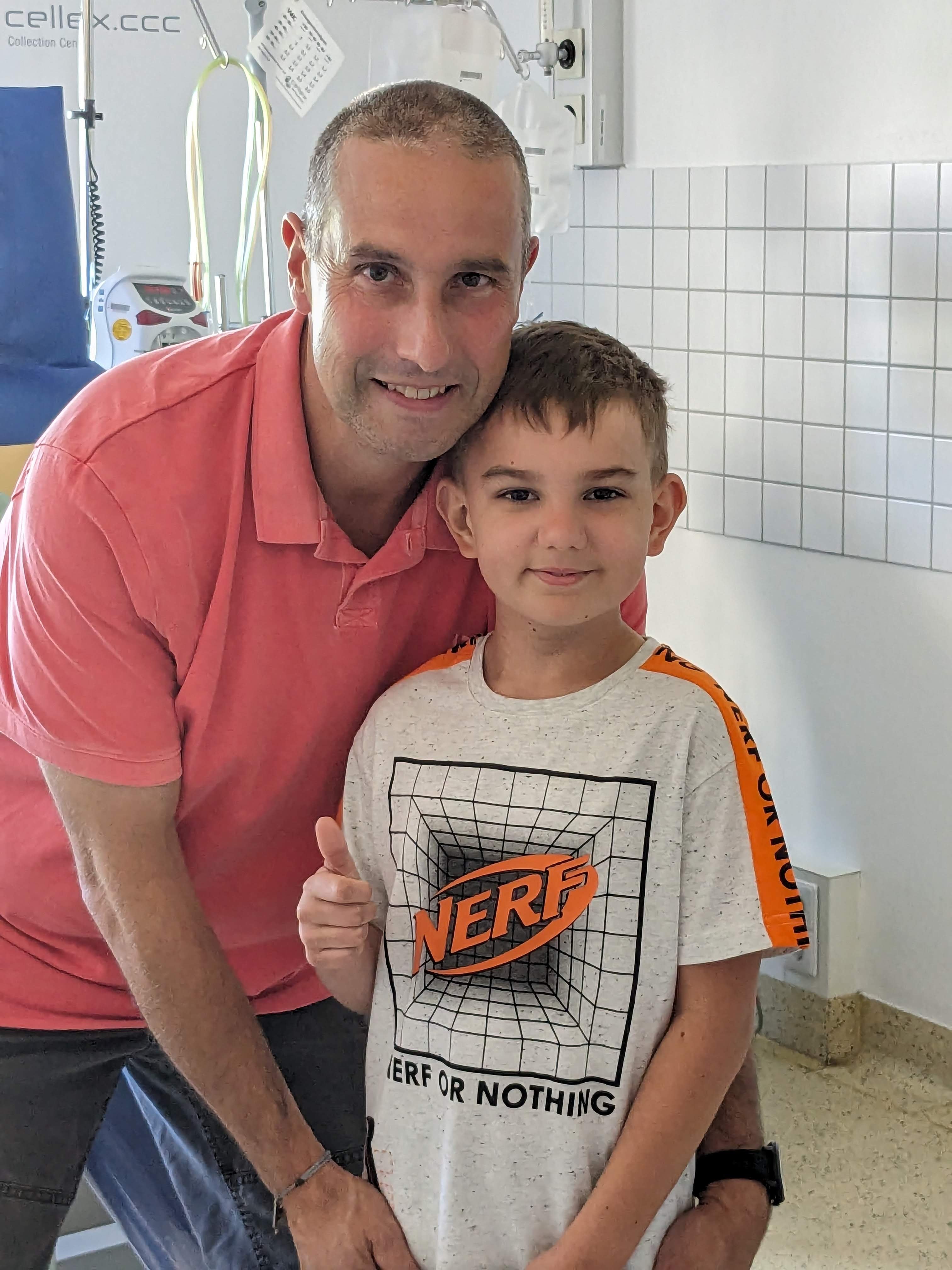 Ein Mann in einem rosa Poloshirt und ein Junge in einem weißen T-Shirt mit „NERF“-Aufdruck stehen nebeneinander in einem Klinikzimmer. Beide lächeln in die Kamera, der Junge zeigt den Daumen hoch. Im Hintergrund sind medizinische Geräte, eine Fliese an der Wand und ein Infusionsständer zu sehen.