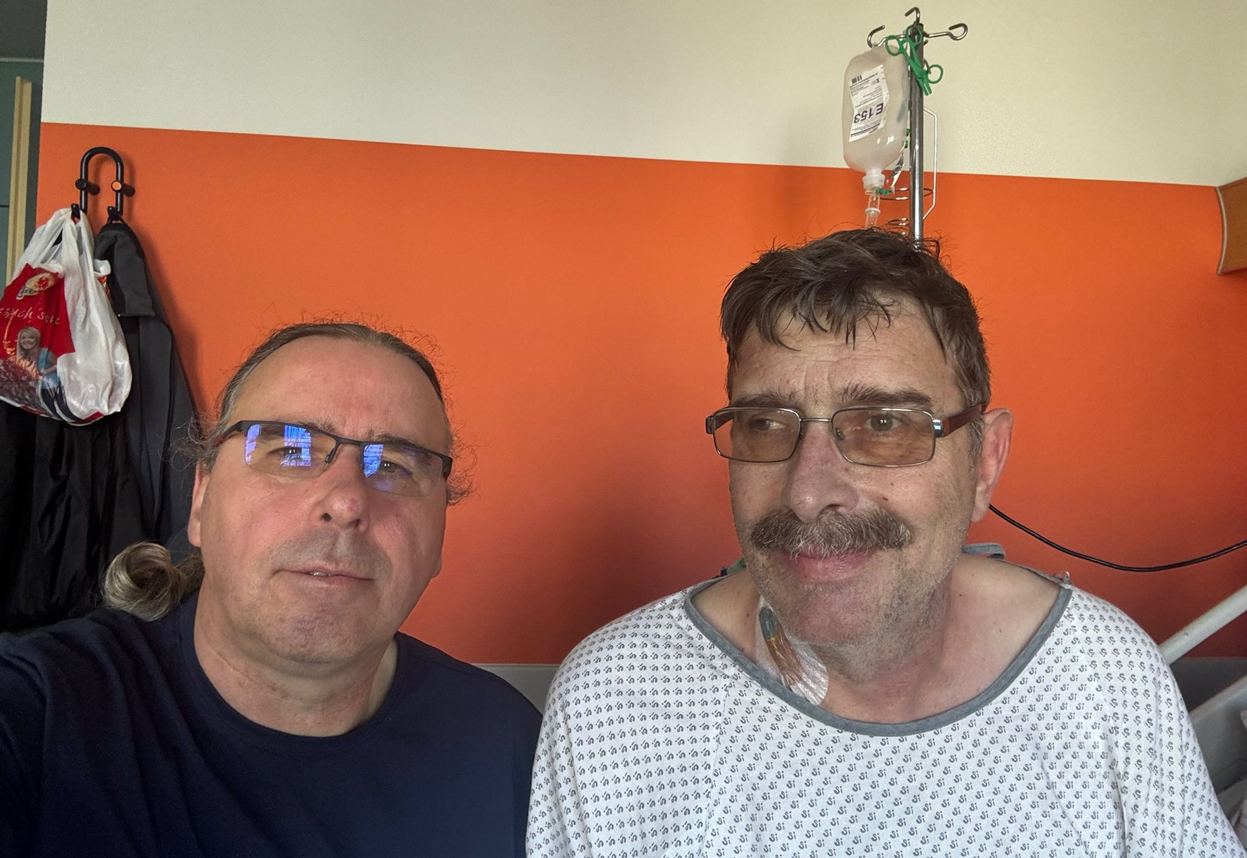 Zwei Männer mit Brille lächelnd vor einer weiß-orangenen Wand.