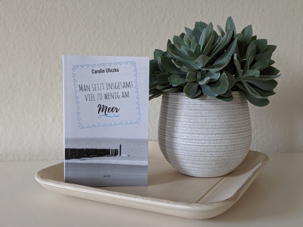 Eine Pflanze in einem Topf auf einem Tablett. Neben der Pflanze steht ein Buch von Carolin Uliczka mit dem Titel "Man sitzt insgesamt viel zu wenig am Meer"