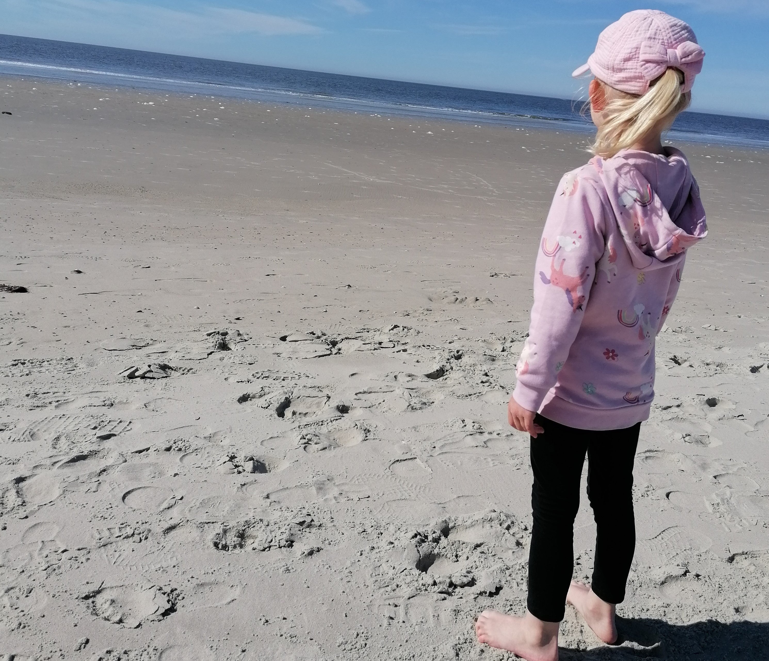Ein blondes Mädchen mit rosa Cap und Hoodie steht mit dem Rücken zur Kamera und schaut aufs Meer.
