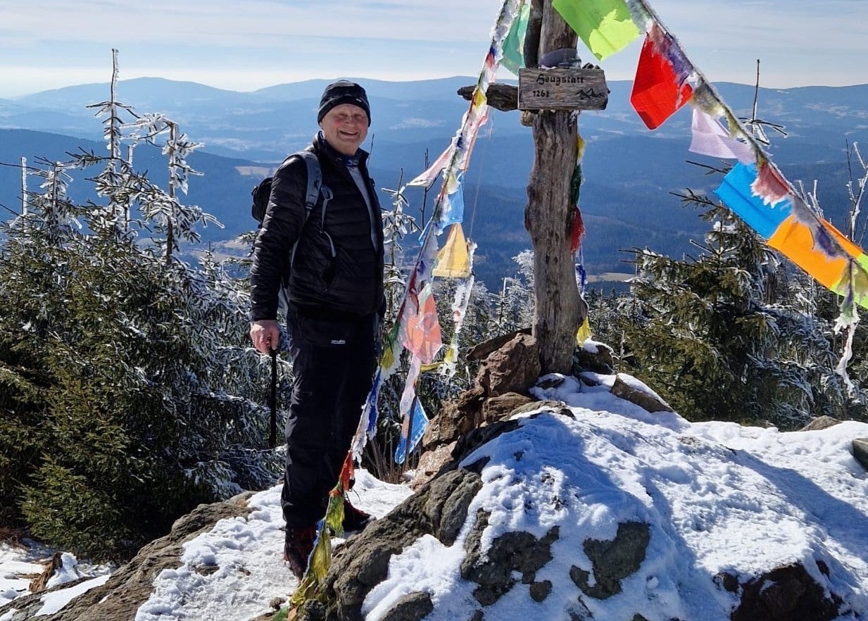 Martin steht neben einem Gipfelkreuz auf einem schneebedeckten Berg und lächelt in die Kamera.