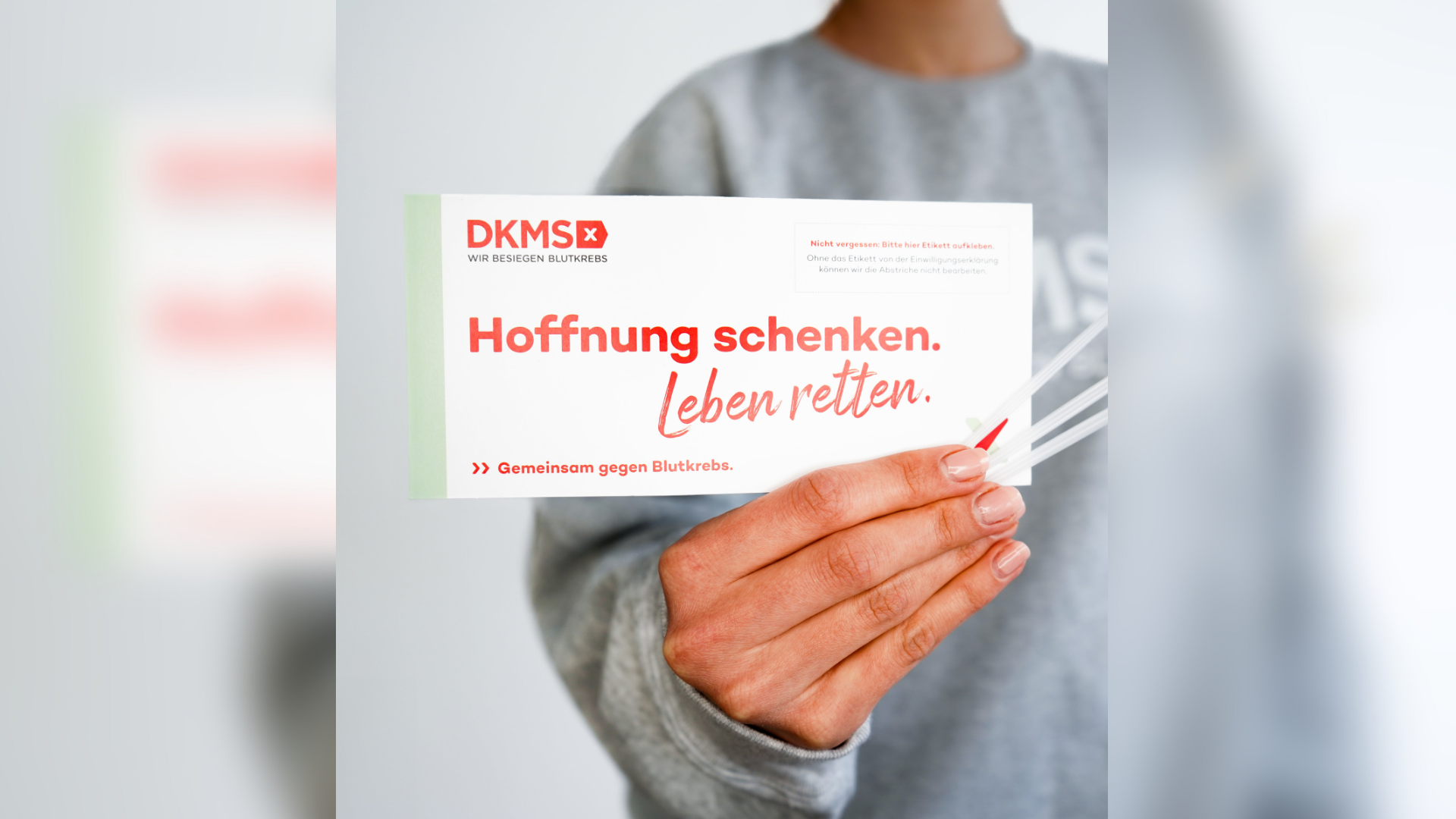 Eine Person trägt einen grauen Pullover und hält das DKMS Registrierungsset in die Kamera. 